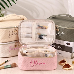 May include: A collection of personalized makeup bags in various colors, including pink, beige, and olive green. Each bag features a woven texture, a top handle, and a zipper closure. The bags are open, revealing makeup brushes and other beauty products.