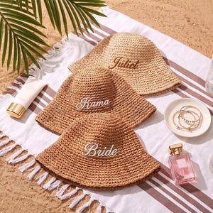 Op de afbeelding: Drie geweven strooien bucket hats met geborduurde tekst. De bovenste hoed is lichtbeige met de naam Juliet in bruin. De middelste hoed is middenbruin met de naam Kama in wit. De onderste hoed is middenbruin met het woord Bride in wit.