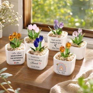 May include: Five white square ceramic planters containing glass tulip flowers in orange, pink, blue, and purple with green stems. Each planter features personalized text, including names like Mrs. Lynn, Hannah, Miss Thea, and Mrs. Taylor, with decorative hearts and a rainbow graphic.
