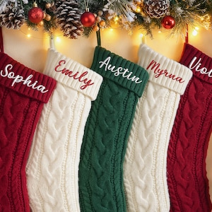 May include: Five cable-knit Christmas stockings in red, white, and green, each with a name embroidered in a contrasting color. The stockings are decorated with festive greenery, ornaments, and lights.