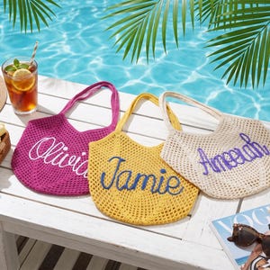 May include: Three crocheted tote bags in fuchsia, yellow, and cream, each with a name embroidered on the front. The bags sit on a white surface near a pool, with a drink, sunglasses, and a magazine.