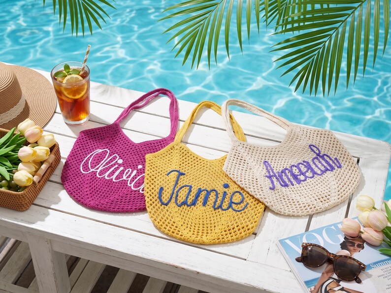 May include: Three crocheted tote bags in fuchsia, yellow, and cream, each with a name embroidered on the front. The bags sit on a white surface near a pool, with a drink, sunglasses, and a magazine.
