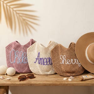 May include: Three open-weave tote bags in pink, cream, and brown, each embroidered with a name in a contrasting color. A wide-brimmed sun hat, sunglasses, and sunscreen are on a wooden table, suggesting a beach theme.