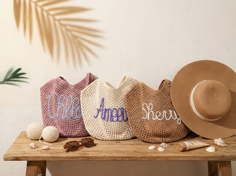 May include: Three open-weave tote bags in pink, cream, and brown, each embroidered with a name in a contrasting color. A wide-brimmed sun hat, sunglasses, and sunscreen are on a wooden table, suggesting a beach theme.