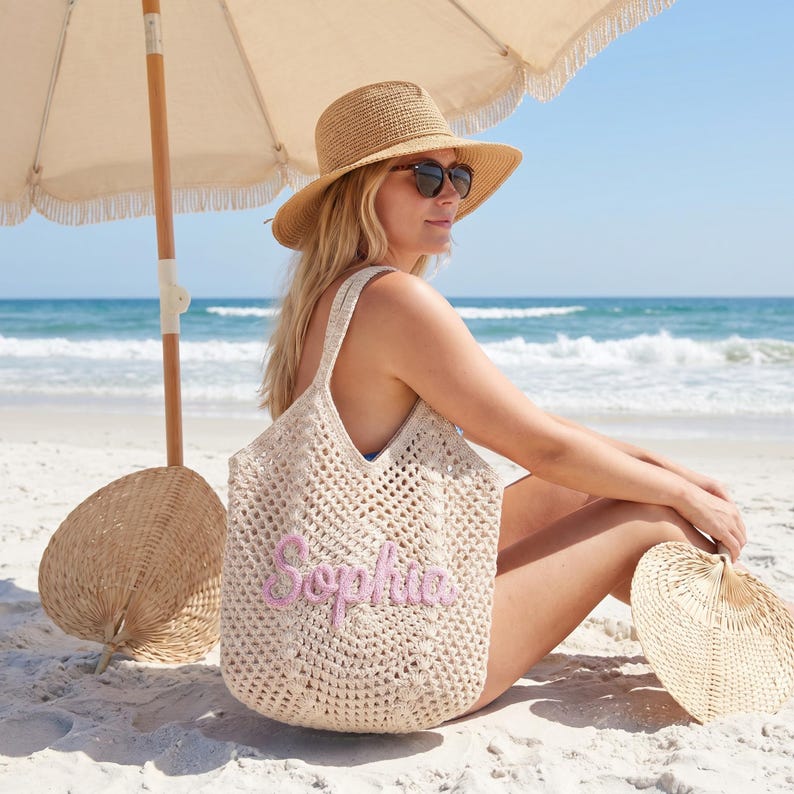 May include: A beige crochet tote bag with the name "Sophia" embroidered in pink. The bag has a wide opening and shoulder straps. A straw hat and sunglasses are also visible. The scene is set on a sandy beach with the ocean in the background.