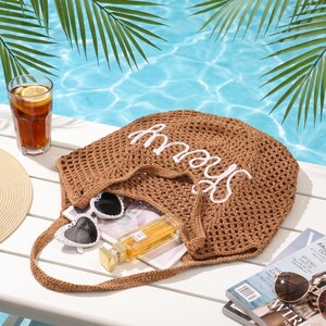 May include: A beach-themed still life featuring a brown crocheted tote bag with the word "Sherry" in white, sunglasses, a straw hat, a glass of iced tea with lemon, and a fashion magazine. Palm fronds and a pool are in the background.