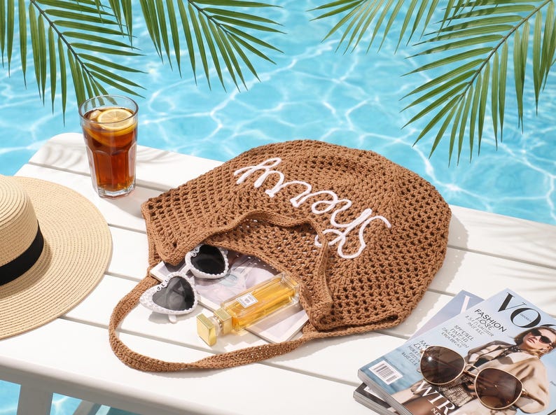 May include: A beach-themed still life featuring a brown crocheted tote bag with the word "Sherry" in white, sunglasses, a straw hat, a glass of iced tea with lemon, and a fashion magazine. Palm fronds and a pool are in the background.