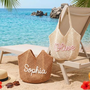 May include: Two woven tote bags, one beige and one brown, sit on a sandy beach. The beige bag has the name "Olive" embroidered in pink, while the brown bag has "Sophia" in white. A straw hat, sunglasses, and a tropical drink are also on the sand.