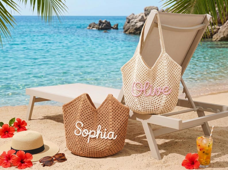 May include: Two woven tote bags, one beige and one brown, sit on a sandy beach. The beige bag has the name "Olive" embroidered in pink, while the brown bag has "Sophia" in white. A straw hat, sunglasses, and a tropical drink are also on the sand.