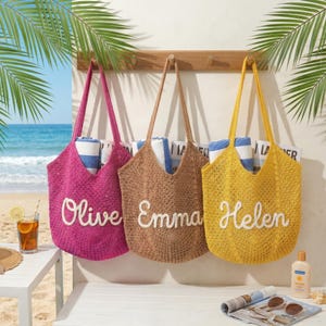 May include: Three crochet tote bags in pink, brown, and yellow hang on a wooden rack. Each bag has a name embroidered in white: Olive, Emma, and Helen. The bags are filled with rolled-up blue and white striped towels, perfect for a day at the beach.