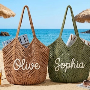 May include: Two open-weave tote bags on a sandy beach, one brown and one olive green, each with a name in white script. The brown bag says "Olive" and the green bag says "Sophia". Both bags contain magazines.