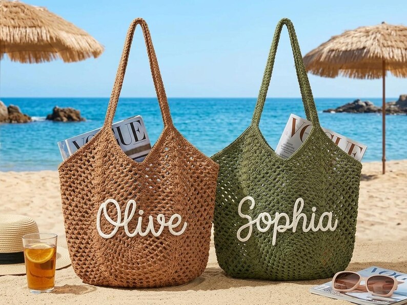 May include: Two open-weave tote bags on a sandy beach, one brown and one olive green, each with a name in white script. The brown bag says "Olive" and the green bag says "Sophia". Both bags contain magazines.