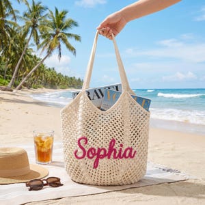 May include: A cream-colored, open-weave tote bag with the name "Sophia" embroidered in pink. The bag is filled with magazines and held by a person on a beach. A straw hat, sunglasses, and a glass of iced tea are also on the beach.
