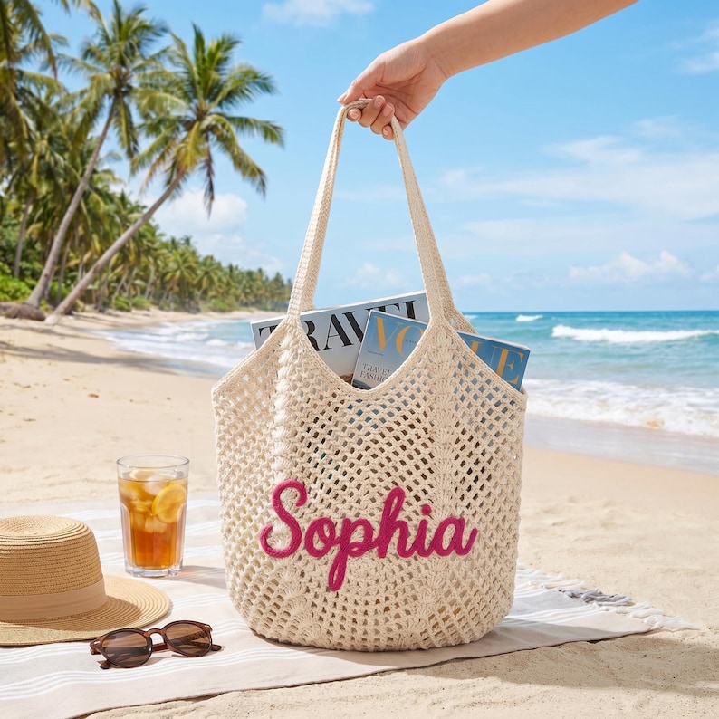 May include: A cream-colored, open-weave tote bag with the name "Sophia" embroidered in pink. The bag is filled with magazines and held by a person on a beach. A straw hat, sunglasses, and a glass of iced tea are also on the beach.