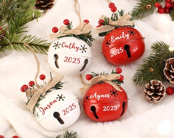 Puede incluir: Cuatro adornos de campana navideños, dos blancos y dos rojos, con nombres y el año 2025. Cada campana tiene un diseño de ojo de cerradura, un lazo y bayas. Decoración festiva navideña.