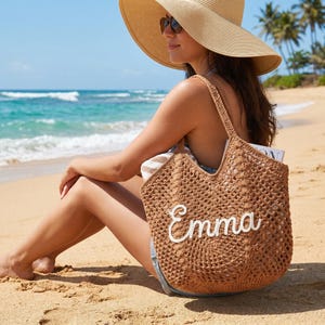 May include: A brown, open-weave beach tote bag with the name "Emma" in white script. The bag is filled with a striped towel and a magazine. The person is wearing a large sun hat and sunglasses, sitting on a sandy beach.