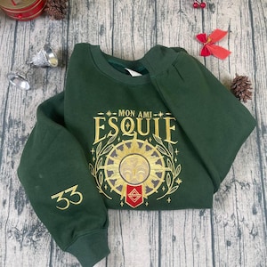 Puede incluir: Una sudadera verde oscuro con el texto "MON AMI ESQUIF" en letras doradas, junto con un diseño de sol. El número "33" está en la manga izquierda. La sudadera está doblada sobre una superficie de madera.