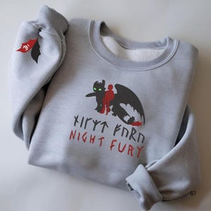 Könnte beinhalten: Hellgraues Sweatshirt mit einem schwarzen und roten Motiv eines Drachen und zweier Figuren, mit dem Text "Night Fury" in Rot und Schwarz. Auf dem linken Ärmel befindet sich ein kleines rotes und schwarzes Emblem. Das Sweatshirt ist gefaltet.