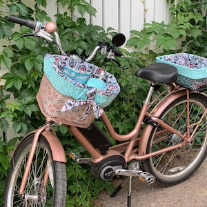 May include: A rose gold electric bicycle with a black seat and two baskets. The front basket is lined with a turquoise and white patterned fabric. The rear basket is lined with a turquoise and black patterned fabric.