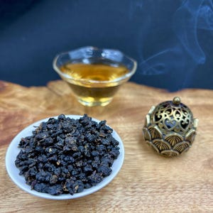May include: A still life featuring tea and incense. A small white bowl holds dark tea leaves, next to a decorative gold incense burner. A glass cup of tea sits in the background, with wisps of smoke rising.