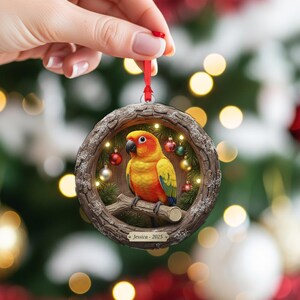 May include: A round Christmas ornament featuring a vibrant yellow and orange parrot perched on a branch within a wooden frame. The frame is adorned with small, colorful ornaments and lights. The ornament is suspended by a red ribbon. The text "Jessica - 2025" is visible.
