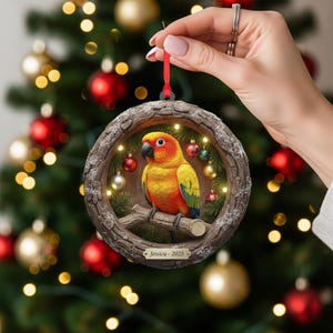 May include: A round Christmas ornament featuring a vibrant yellow, orange, and green parrot perched on a branch. The ornament is framed by a wooden-like border with small lights and miniature ornaments. The text "Jessica - 2025" is displayed on the bottom.