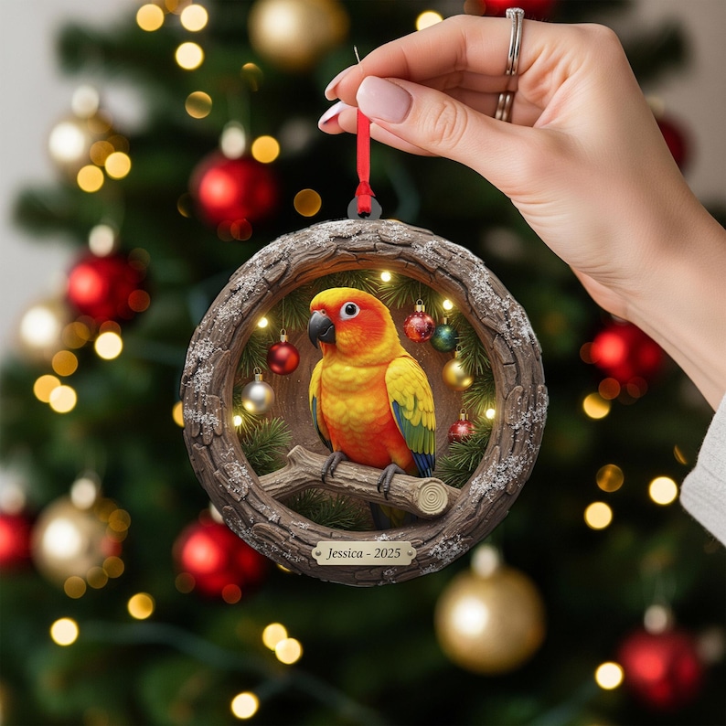 May include: A round Christmas ornament featuring a vibrant yellow, orange, and green parrot perched on a branch. The ornament is framed by a wooden-like border with small lights and miniature ornaments. The text "Jessica - 2025" is displayed on the bottom.