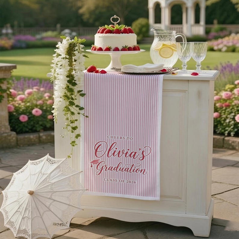 May include: A white cake with strawberries sits on a white pedestal, next to a pitcher of lemonade and two glasses. A pink and white striped table runner reads "Cheers to Olivia's Graduation Class of 2026."