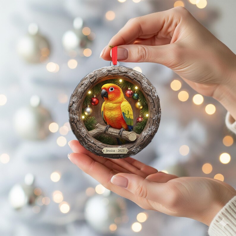 May include: A round Christmas ornament featuring a colorful parrot perched on a branch inside a wooden frame. The ornament has a red ribbon for hanging and is decorated with small lights and miniature ornaments. The name "Jessica - 2025" is printed on the bottom.