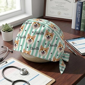 May include: A medical scrub cap featuring a repeating pattern of smiling corgi faces, paw prints, and the name "Charlie" in a script font. The background has vertical stripes in white and light green.