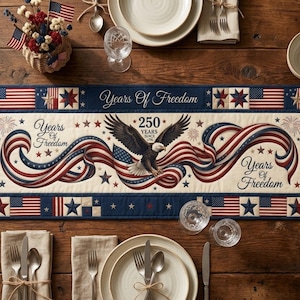 Puede incluir: Un camino de mesa patriótico con un diseño que presenta la bandera estadounidense, estrellas y un águila. Se muestra el texto "Years Of Freedom" y "250 Years Since 1776". El camino de mesa está colocado sobre una mesa de madera con cubiertos y elementos decorativos.