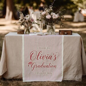 May include: A table setting with a pink and white striped graduation table runner. The runner reads "Cheers to Olivia's Graduation Class of 2026" in a script font. The table is set with flowers and small decorative boxes.