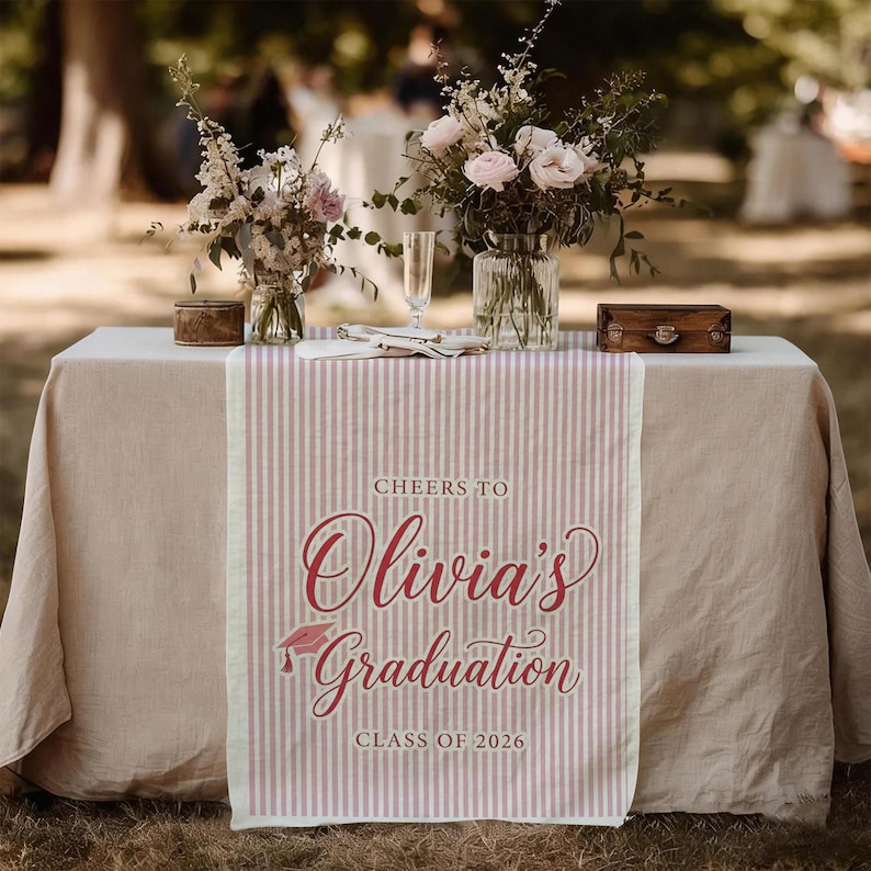 May include: A table setting with a pink and white striped graduation table runner. The runner reads "Cheers to Olivia's Graduation Class of 2026" in a script font. The table is set with flowers and small decorative boxes.