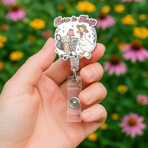 May include: A clear badge reel featuring a cartoon goose pushing a shopping trolley filled with thrifted items. The text "Born to Thrift" is above the goose. The reel has a clear plastic strap and a metal button.