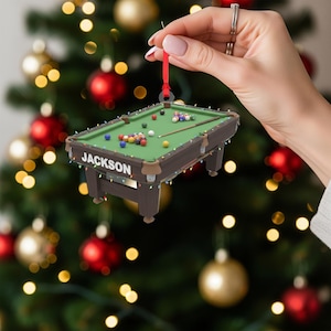 May include: A miniature pool table Christmas ornament with the name "Jackson" on the side. The ornament features a green felt surface, a cue stick, and a collection of billiard balls. It is suspended by a red ribbon.