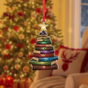 May include: A Christmas tree ornament shaped like a stack of colorful books, topped with a gold star. The books are various colors, including green, red, and blue, and are suspended by a red ribbon. The background features a blurred Christmas tree.