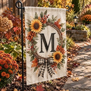 May include: A rectangular garden flag featuring a white wood-grain background with a central wreath of sunflowers, autumn leaves, and berries. A large black letter M is centred within the wreath, which is accented by a black and white buffalo check bow.