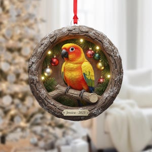 May include: A round Christmas ornament featuring a vibrant yellow, orange, and green parrot perched on a branch within a wooden frame. The frame is decorated with small Christmas lights, ornaments, and the text "Jessica - 2025". A red ribbon is attached for hanging.