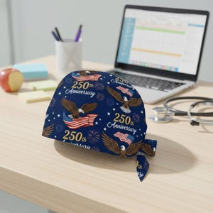 May include: A navy blue scrub cap featuring a repeating pattern of eagles, American flags, and the text "250th Anniversary." The design includes white stars and fireworks. The cap is tied at the back.