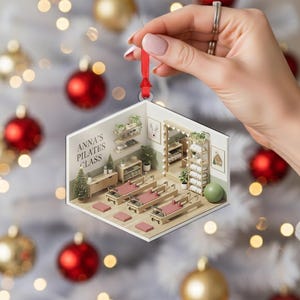 May include: A Pilates class Christmas ornament. The ornament features a miniature Pilates studio with exercise equipment, plants, and the text "ANNA'S PILATES CLASS". The ornament is held by a hand with a red ribbon.