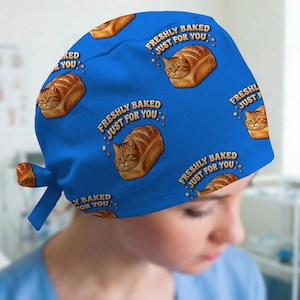 May include: A blue scrub cap featuring a repeating pattern of orange tabby cats shaped like loaves of bread. Each cat illustration is accompanied by the text "FRESHLY BAKED JUST FOR YOU" in a curved brown font, accented with small yellow stars.