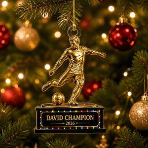 May include: A gold-toned soccer player figurine ornament suspended by a gold string. The figure is in a kicking motion with a soccer ball on a black rectangular base. The base features the text DAVID CHAMPION 2026 with small colorful lights.