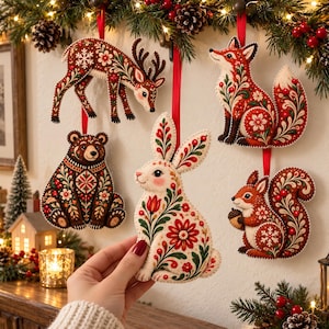 May include: A set of five felt animal ornaments featuring a deer, fox, bear, rabbit, and squirrel. Each ornament is decorated with intricate red, green, and white floral patterns and hangs from a red ribbon against a white wall.