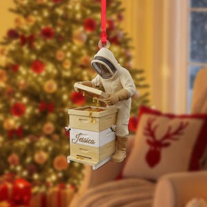 May include: A Christmas tree ornament depicting a beekeeper in a protective suit examining a wooden beehive. The ornament hangs from a red ribbon. The beehive is labelled "Jessica". The background is a blurred Christmas tree.