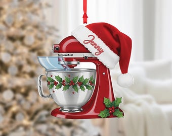 May include: A red KitchenAid mixer Christmas ornament with a Santa hat that says "Jenny". The mixer has a silver bowl decorated with holly and berries. A red ribbon is attached for hanging. Festive holiday decor.