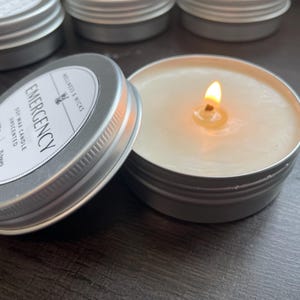 May include: A lit, unscented soy wax candle in a silver tin with the word "EMERGENCY" on the label. The label also includes the text "Wellness & Wicks" and "Long lasting clean burn". Several similar candles are visible.