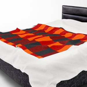 May include: A patterned throw blanket in shades of orange, red, and black, arranged in a geometric design. The blanket is placed on a bed with a white sheet and a black headboard. The blanket adds a vibrant touch to the bedroom decor.