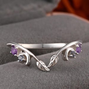 May include: A silver-colored ring with a leaf and vine design. The ring features two purple and two gray gemstones. The ring is set against a gray background.