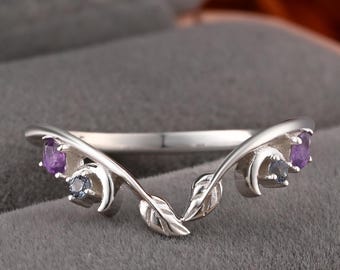 May include: A silver-colored ring with a leaf and vine design. The ring features two purple and two gray gemstones. The ring is set against a gray background.