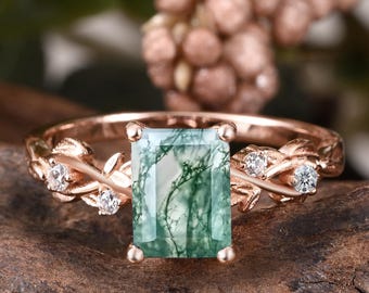 May include: Rose gold ring featuring a rectangular, green moss agate gemstone. The band has a leaf and vine design with small, clear accent stones. The ring is set against a natural wood background.